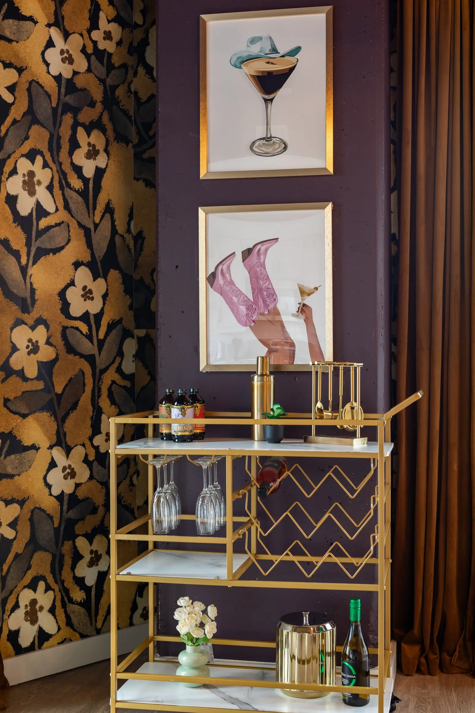 Gold bar cart styled with glassware against plum walls and gold floral wallcovering, beneath a cocktail print and a pink cowboy boot print