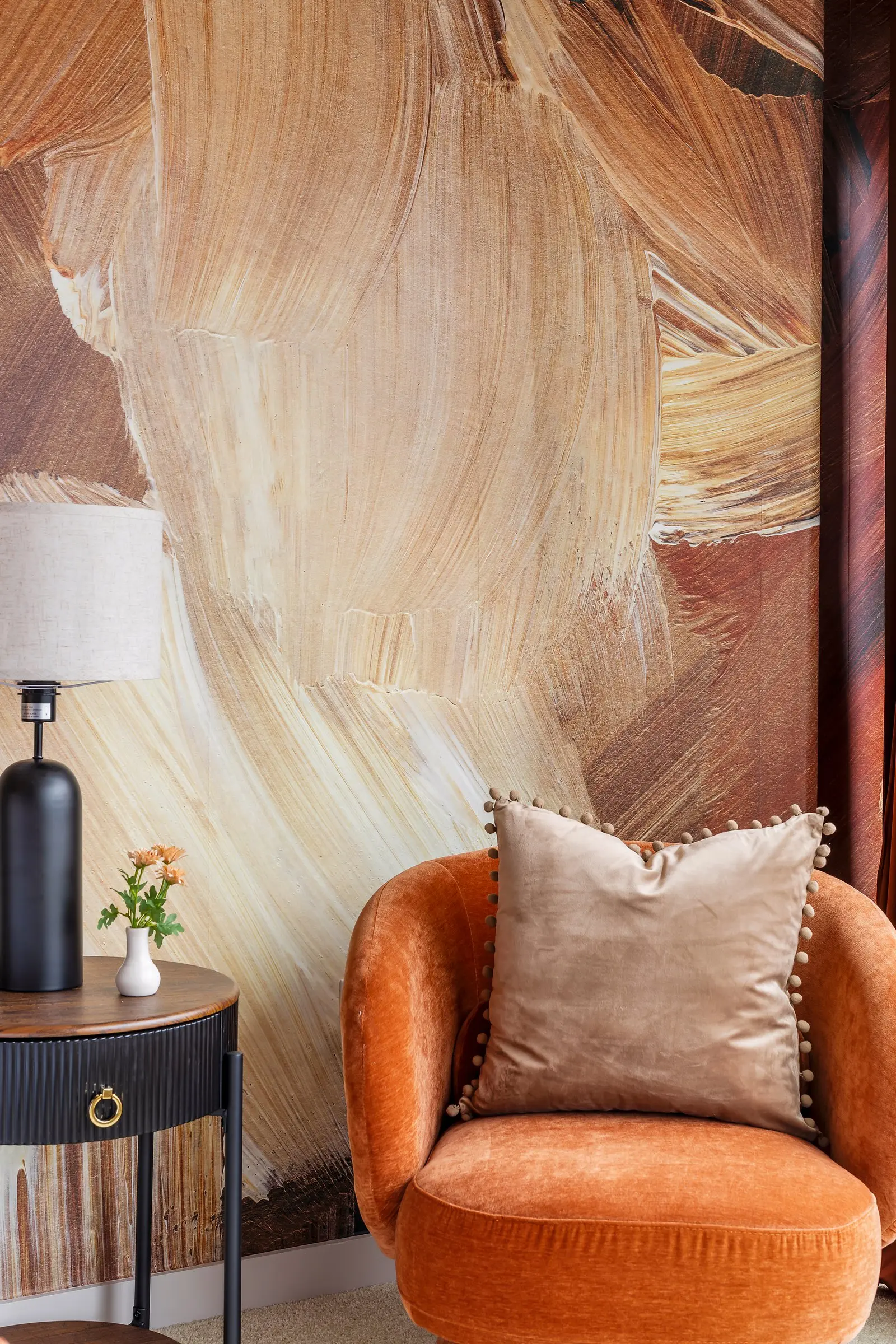 Burnt orange sculptural chair beside a black side table and lamp against a painterly brushstroke mural