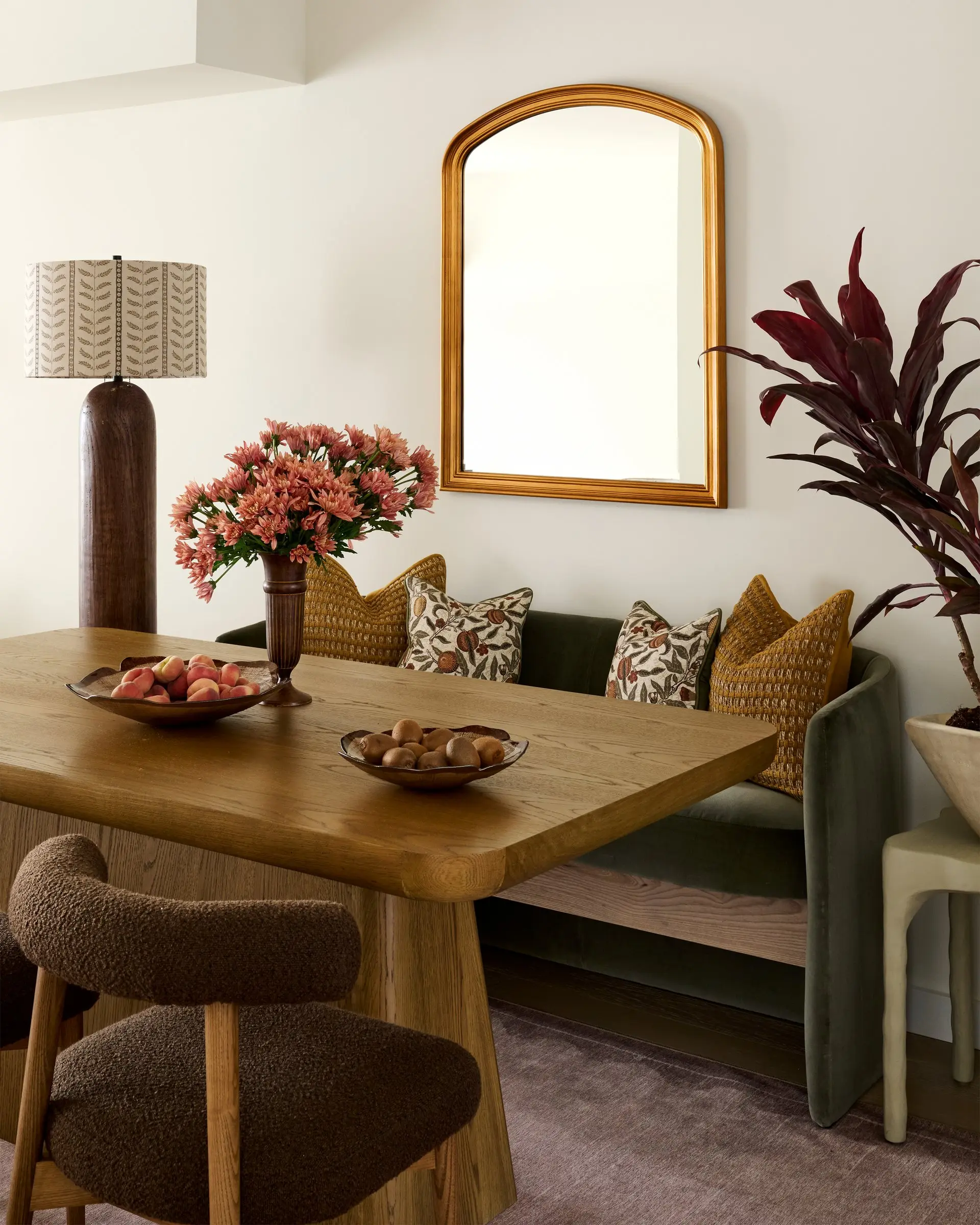 Olive velvet banquette with a sculptural wood dining table, boucle chairs, an arched brass mirror and coral blooms