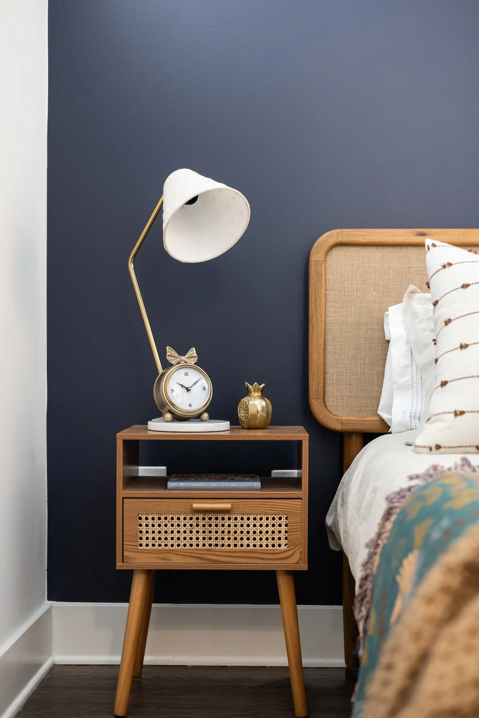 Cane nightstand with a brass clock and an arched task lamp against a navy wall