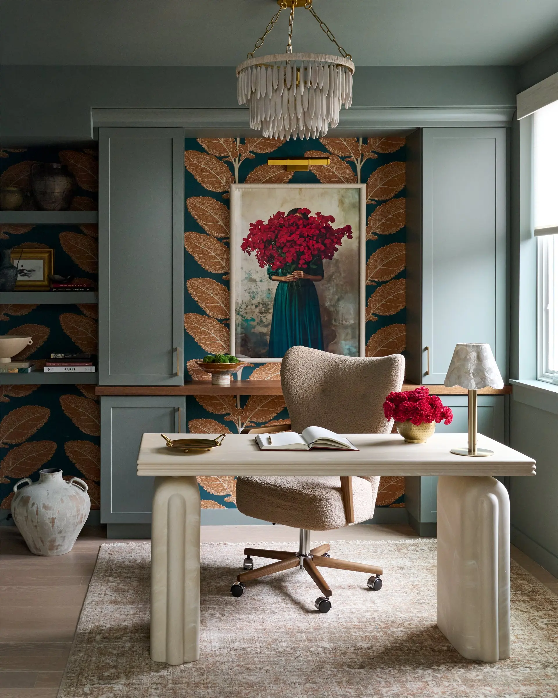 Home study with teal-gray built-ins, botanical wallcovering, a crimson floral portrait, a plaster desk and a tiered chandelier