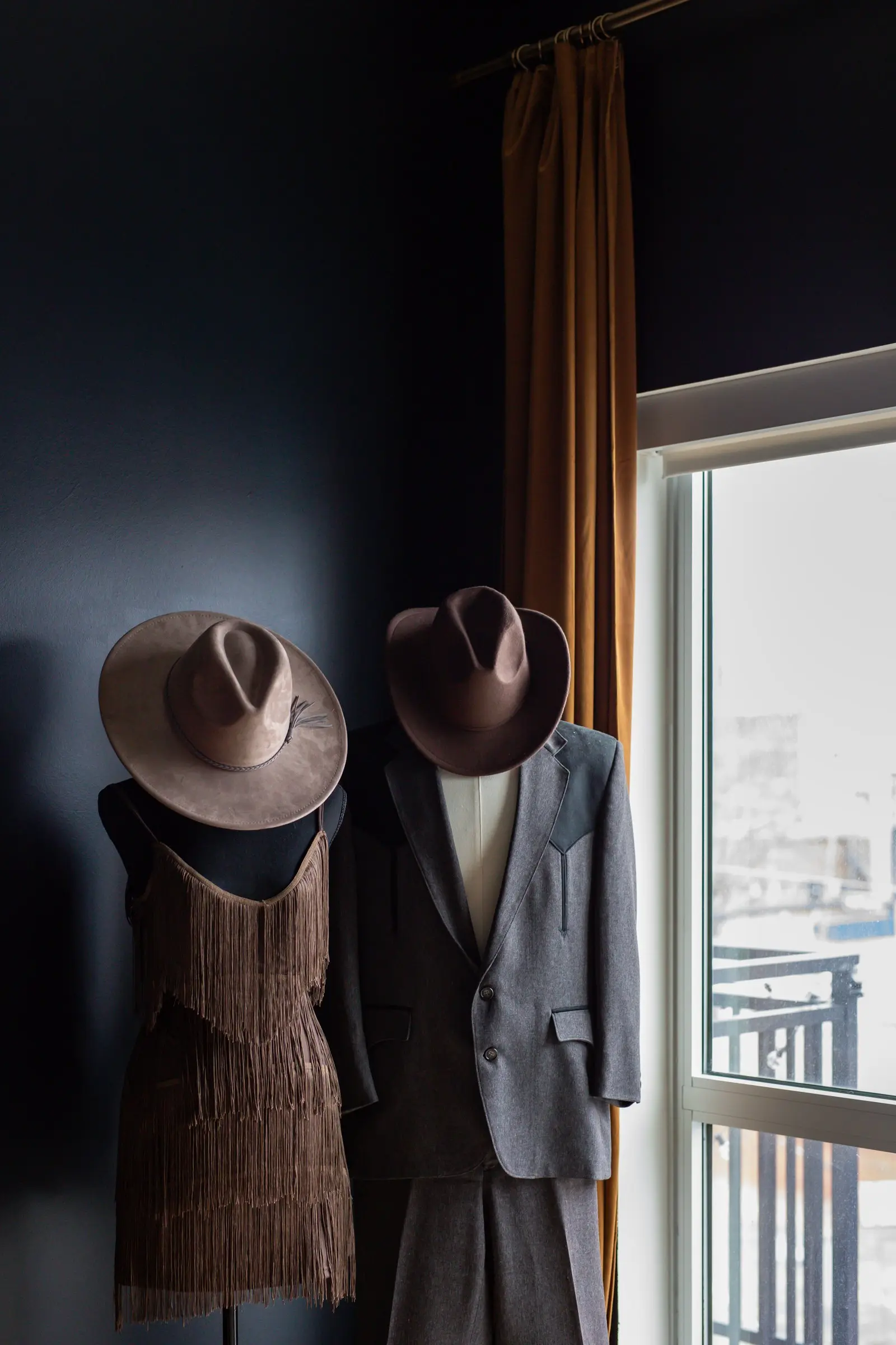 Western outfits and hats on dress forms against a deep navy wall beside the window